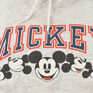 Op de afbeelding: Een lichtgrijze gem&ecirc;leerde hoodie met het woord MICKEY in grote marineblauwe letters met een rode omlijning. Onder de tekst staat een afbeelding van vijf zwart-witte Mickey Mouse-hoofden op een rij, voorzien van witte trekkoorden.