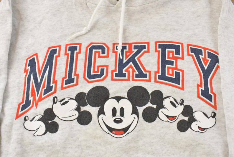 Op de afbeelding: Een lichtgrijze gem&ecirc;leerde hoodie met het woord MICKEY in grote marineblauwe letters met een rode omlijning. Onder de tekst staat een afbeelding van vijf zwart-witte Mickey Mouse-hoofden op een rij, voorzien van witte trekkoorden.