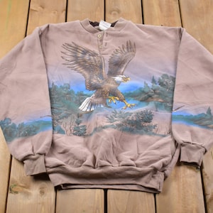 Peut inclure: Un sweat-shirt beige avec un motif détaillé d'un aigle à tête blanche en vol au-dessus d'un paysage avec des arbres et un plan d'eau. L'aigle a des plumes marron et blanches. Le sweat-shirt a un col boutonné et des manches longues.