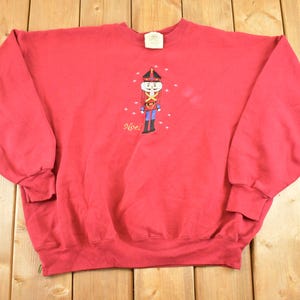May include: A red sweatshirt with a crew neck and long sleeves. The front features an embroidered cartoon nutcracker soldier with the word "Noel" in gold script. The sweatshirt is made of a soft material.