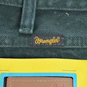 May include: Close-up of a dark green pair of Wrangler jeans with a yellow Wrangler label sewn onto the back pocket. The label has the word "Wrangler" in gold lettering. A yellow tag with a blue border and the words "True Performance" is visible in the bottom right corner of the image.