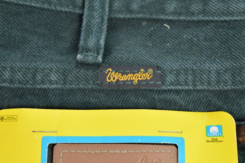 May include: Close-up of a dark green pair of Wrangler jeans with a yellow Wrangler label sewn onto the back pocket. The label has the word "Wrangler" in gold lettering. A yellow tag with a blue border and the words "True Performance" is visible in the bottom right corner of the image.