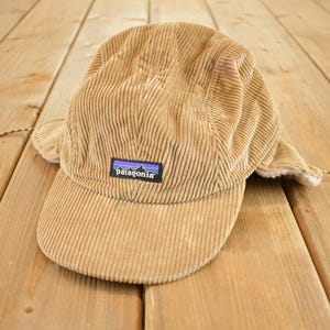 Può includere: Cappellino in velluto a coste beige con paraorecchie e piccola visiera. Il cappellino presenta una toppa rettangolare con il marchio "Patagonia" e un motivo montano. Ha un cordino.