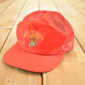 Op de afbeelding: Rode truckerpet met een mesh achterkant en een gebogen klep. De pet heeft de tekst "MEDAL OF HONOR GOLF COURSE QUANTICO" in gouden letters, samen met een embleem. De pet is gemaakt van een zacht materiaal.