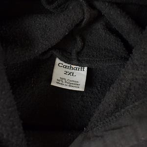 May include: Close-up of a black Carhartt hoodie. The tag reads "Carhartt 2XL" with fabric composition details: 50% cotton, 50% polyester. Made in Mexico.