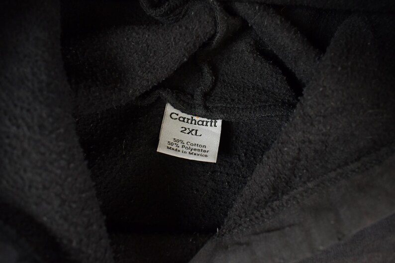 May include: Close-up of a black Carhartt hoodie. The tag reads "Carhartt 2XL" with fabric composition details: 50% cotton, 50% polyester. Made in Mexico.