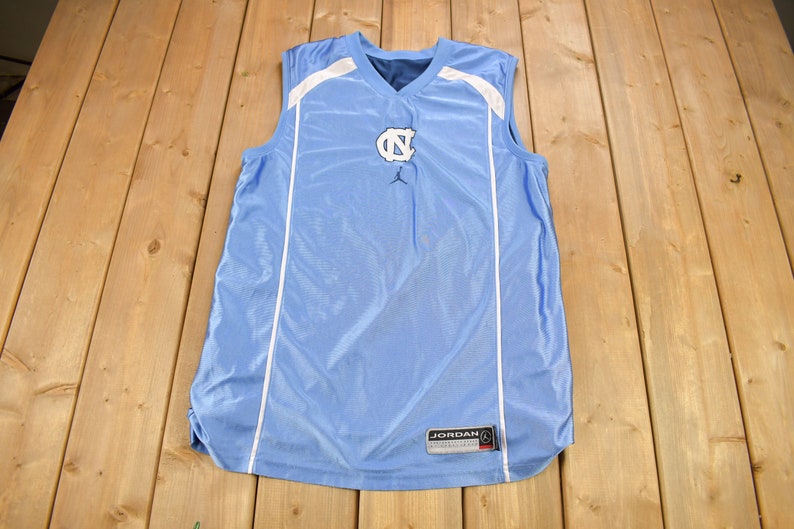 May include: A light blue sleeveless basketball jersey with white trim and the letters "NC" in navy blue on the front. The Jordan logo is below the letters. The jersey has a white tag on the bottom right side that says "JORDAN" in black.