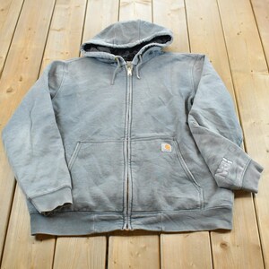 Peut inclure: Un sweat-shirt à capuche zippé gris clair doté d'une poche kangourou à l'avant, d'une capuche à cordon avec une doublure intérieure foncée et d'une petite étiquette rectangulaire de la marque Carhartt sur la poche. Le poignet gauche porte du texte brodé.