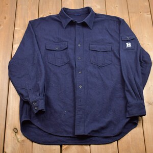 Può includere: Una camicia abbottonata a maniche lunghe blu navy con due tasche a patta e una toppa sulla manica sinistra che dice "B".