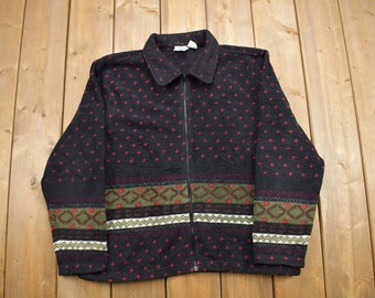 Könnte beinhalten: Eine schwarze Fleecejacke mit einem roten und grünen geometrischen Muster. Die Jacke hat einen Reißverschluss und einen Kragen.