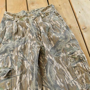 May include: Camouflage cargo pants in shades of brown, green, and gray. The pants feature multiple pockets and a button closure. The fabric has a textured appearance, and the pants are laid out on a wooden surface.