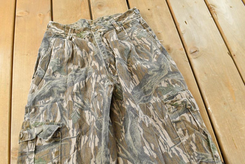 May include: Camouflage cargo pants in shades of brown, green, and gray. The pants feature multiple pockets and a button closure. The fabric has a textured appearance, and the pants are laid out on a wooden surface.