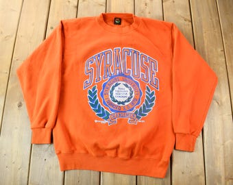 May include: Orange crew neck sweatshirt with the word "Syracuse" in blue and white lettering. The sweatshirt features a university emblem with text and a laurel wreath design. The text "Founded A.D. 1870" is also visible.