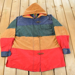 May include: A colorful hooded jacket with a patchwork design. The jacket features a color block pattern with red, tan, green, and blue sections. It has toggle closures and a pocket on the chest. The hood is tan.