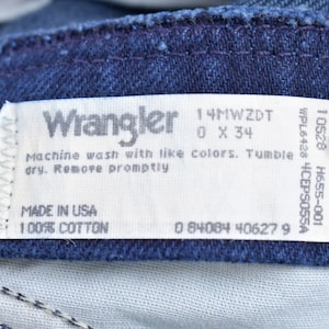 May include: A white Wrangler jeans care label with the text "Wrangler", "14MWZDT", "0 X 34", "Machine wash with like colors. Tumble dry. Remove promptly", "MADE IN USA", "100% COTTON", "0 84084 406279", "10528 H655-001", "WPL6428 4CEPS055A".