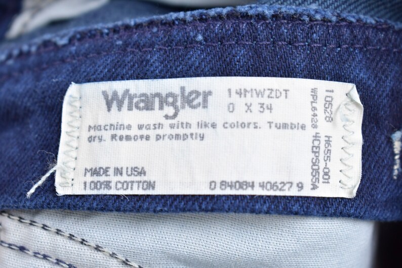 May include: A white Wrangler jeans care label with the text "Wrangler", "14MWZDT", "0 X 34", "Machine wash with like colors. Tumble dry. Remove promptly", "MADE IN USA", "100% COTTON", "0 84084 406279", "10528 H655-001", "WPL6428 4CEPS055A".