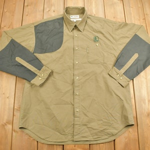 May include: A long-sleeved, tan button-down shirt with dark green and gray fabric panels on the shoulders and sleeves. The shirt has a Columbia logo on the left chest.