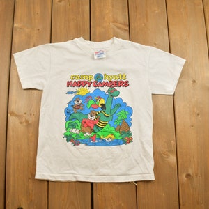 Könnte beinhalten: Weißes T-Shirt mit einem Cartoon-Motiv eines grünen Dinosauriers, eines Eichhörnchens und eines Vogels. Der Text "camp hyatt happy campers" ist auf das Shirt gedruckt.