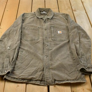 May include: A tan long-sleeve button-up shirt featuring a classic collar, two chest pockets, and a small rectangular Carhartt brand patch on the left chest pocket. The shirt has metal snap buttons down the front and at the sleeve cuffs.