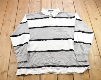 Könnte beinhalten: Ein langärmliges, gestreiftes Rugby-Shirt mit einem grau-weiß gestreiften Muster und schwarzen Streifen. Das Shirt hat ein kleines Logo auf der Brust.