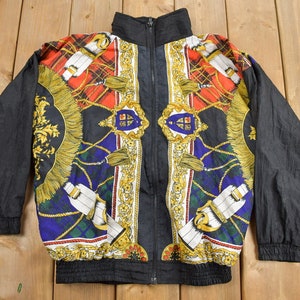 May include: A vintage black zip-up jacket with a high collar. The jacket features a colorful pattern with red plaid, gold accents, and blue and green geometric designs. The sleeves are solid black.
