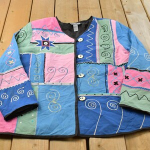 May include: A long-sleeve jacket featuring a patchwork design in shades of blue, pink, and green. The garment includes embroidered spiral, zigzag, and star patterns, a V-neckline, and four round buttons down the front center with black trim.
