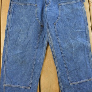 K&ouml;nnte beinhalten: Blaue Jeans-Arbeitshose mit verst&auml;rkten Kniepatches. Die Hose hat ein klassisches F&uuml;nf-Taschen-Design und einen geraden Beinverlauf. Der Denim hat ein verwaschenes Aussehen, was auf Abnutzung und Waschen hindeutet. Die Hose liegt flach auf einer Holzoberfl&auml;che.