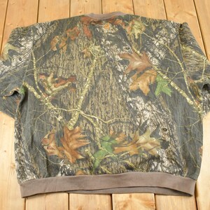 May include: A camouflage-patterned sweatshirt with a brown collar and cuffs. The design features a mix of brown, green, and grey tones, with leaf and tree branch details. The sweatshirt is made of a soft material.