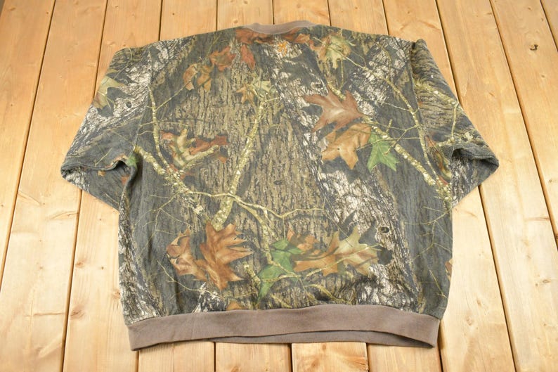 May include: A camouflage-patterned sweatshirt with a brown collar and cuffs. The design features a mix of brown, green, and grey tones, with leaf and tree branch details. The sweatshirt is made of a soft material.