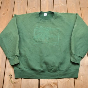 Pode incluir: Um moletom verde com gola redonda e mangas compridas. A frente apresenta um gráfico retangular com a palavra "Montana" e um desenho de paisagem em um tom de verde mais claro. O moletom está estendido sobre uma superfície de madeira.