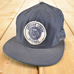 Könnte beinhalten: Marineblaue Baseballkappe mit Mesh-Rückseite und gebogenem Schirm. Die Kappe hat einen runden Aufnäher mit der Aufschrift "AMERICAN LEGION BASEBALL". Die Unterseite des Schirms ist grün.