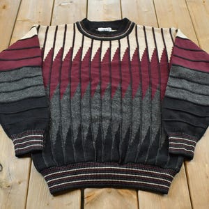 Peut inclure: Un pull en tricot à manches longues avec un col rond et un motif géométrique dans des tons bordeaux, crème, gris anthracite et noir. Le design présente des textures côtelées verticales et des rayures horizontales sur les manches et poignets.