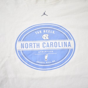 May include: White t-shirt with a blue and white circular logo that says "Tar Heels. North Carolina Athletics Chapel Hill, NC". The logo also features a silhouette of a basketball player and the University of North Carolina Tar Heels logo.