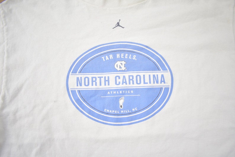 May include: White t-shirt with a blue and white circular logo that says "Tar Heels. North Carolina Athletics Chapel Hill, NC". The logo also features a silhouette of a basketball player and the University of North Carolina Tar Heels logo.