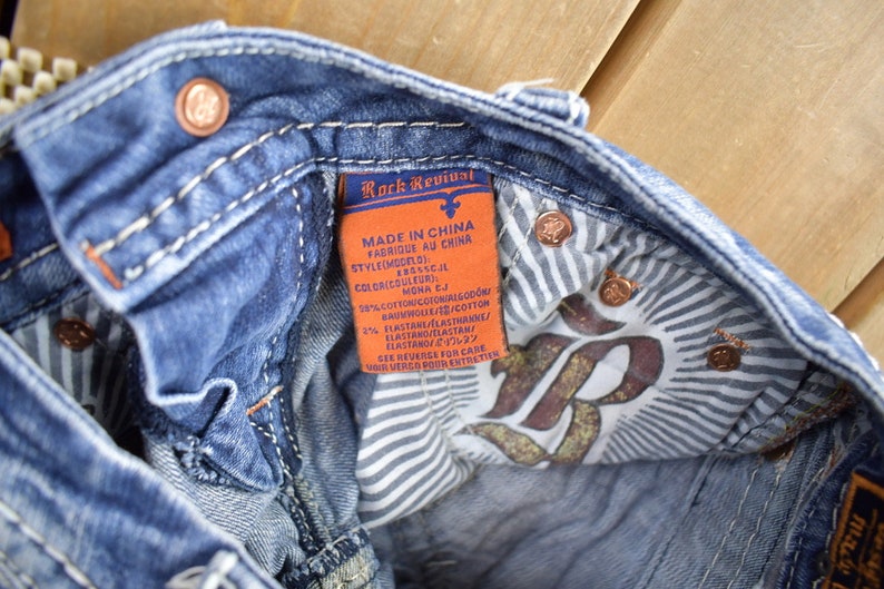K&ouml;nnte beinhalten: Eine Nahaufnahme des Innenfutters einer blauen Jeans. Die Jeans hat ein wei&szlig;-blaues gestreiftes Futter und ein braunes Leder-Etikett mit dem Text "Rock Revival" und Pflegehinweisen in Englisch, Franz&ouml;sisch und Japanisch.
