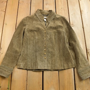 Peut inclure: Une veste en daim marron avec un devant boutonné et des manches longues. La veste a un col et est posée à plat sur une surface en bois.