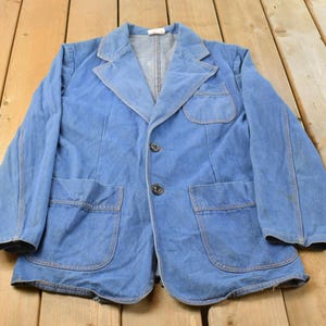 Könnte beinhalten: Ein hellblauer Jeansblazer mit steigendem Revers, zwei dunklen Knöpfen als Verschluss, einer Brusttasche und zwei großen aufgesetzten Taschen an der Taille. Die Jacke hat lange Ärmel und sichtbare Nähte entlang der Kanten und Taschen.