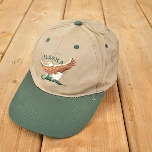 Op de afbeelding: Beige baseballpet met een groene klep. De pet heeft een borduursel van een adelaar met het woord "Alaska". Gemaakt van duurzaam materiaal, met een gebogen klep.