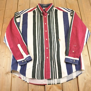 Puede incluir: Camisa de manga larga con un diseño de rayas verticales en rojo, blanco, azul marino y verde. La camisa tiene cuello abotonado, bolsillo en el pecho y puños abotonados. El logo de Tommy Hilfiger está bordado en el pecho.