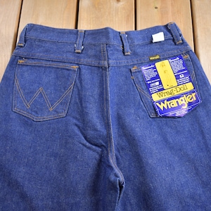 Op de afbeelding: Een donkerblauwe spijkerbroek met een achterzak met een gestikt 'W'-logo. De broek heeft een label met de tekst 'Wrangler' en 'Wrag-Doll'.
