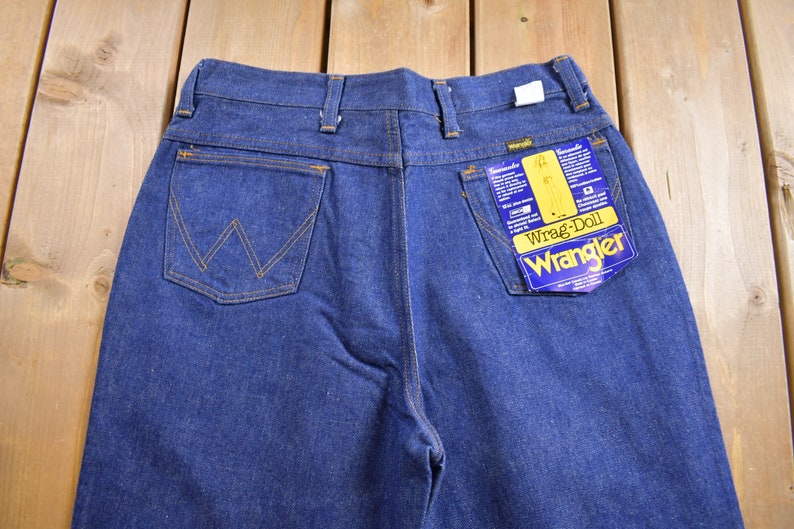 Op de afbeelding: Een donkerblauwe spijkerbroek met een achterzak met een gestikt 'W'-logo. De broek heeft een label met de tekst 'Wrangler' en 'Wrag-Doll'.