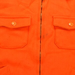 K&ouml;nnte beinhalten: Eine leuchtend orangefarbene Jacke mit Rei&szlig;verschluss und zwei Pattentaschen mit Kn&ouml;pfen. Die Jacke ist aus einem dicken, strukturierten Stoff gefertigt.