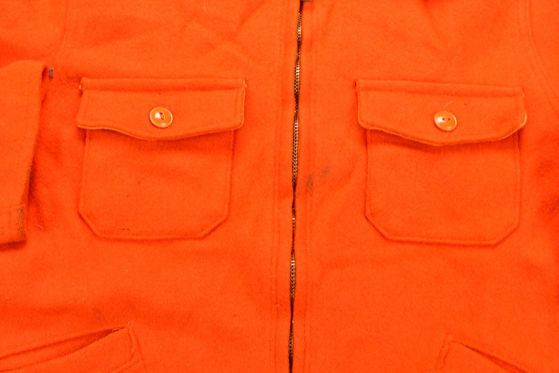 K&ouml;nnte beinhalten: Eine leuchtend orangefarbene Jacke mit Rei&szlig;verschluss und zwei Pattentaschen mit Kn&ouml;pfen. Die Jacke ist aus einem dicken, strukturierten Stoff gefertigt.