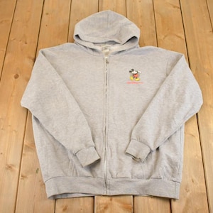 Op de afbeelding: Een lichtgrijze hoodie met ritssluiting, capuchon en lange mouwen. De hoodie heeft een kleine geborduurde afbeelding van Mickey Mouse met tekst eronder. De hoodie is gemaakt van een zacht, comfortabel materiaal.