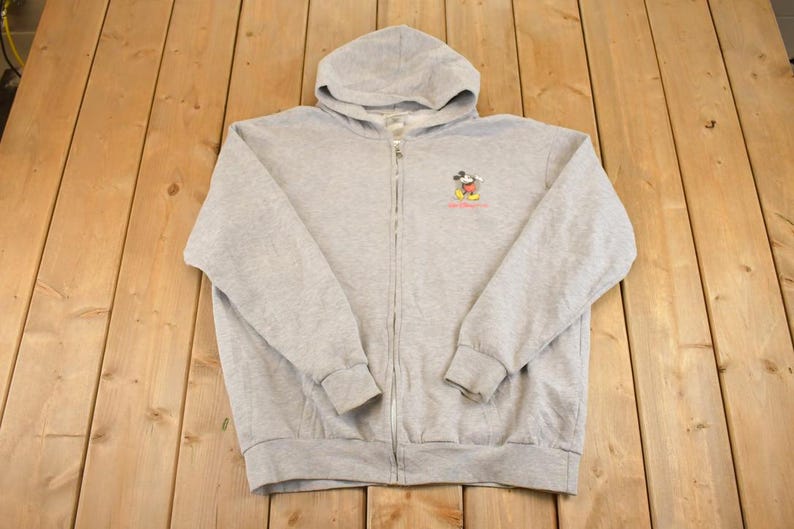 Op de afbeelding: Een lichtgrijze hoodie met ritssluiting, capuchon en lange mouwen. De hoodie heeft een kleine geborduurde afbeelding van Mickey Mouse met tekst eronder. De hoodie is gemaakt van een zacht, comfortabel materiaal.