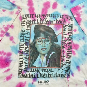 May include: A tie-dye t-shirt with a framed image of a woman wearing a black beret and glasses. The text "Luxottica Eyewear" is printed at the bottom of the image.