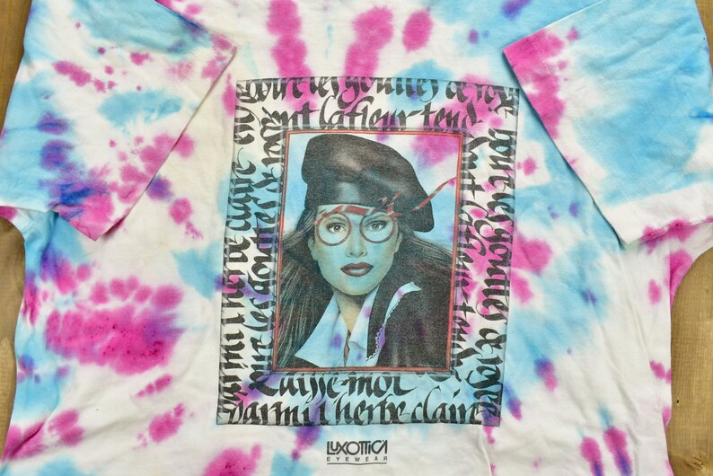 May include: A tie-dye t-shirt with a framed image of a woman wearing a black beret and glasses. The text "Luxottica Eyewear" is printed at the bottom of the image.