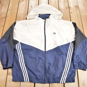 May include: A vintage Adidas hooded jacket in white, navy blue, and black. The jacket features a full zip, the Adidas logo, and three white stripes down each sleeve. The hood is white, and the jacket is laid flat on a wooden surface.