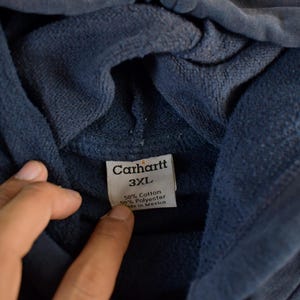 K&ouml;nnte beinhalten: Nahaufnahme eines marineblauen Carhartt-Hoodies. Das Etikett lautet "Carhartt 3XL" und gibt an, dass das Material aus 50 % Baumwolle und 50 % Polyester besteht, hergestellt in Mexiko. Der Stoff des Hoodies wirkt weich und strukturiert.