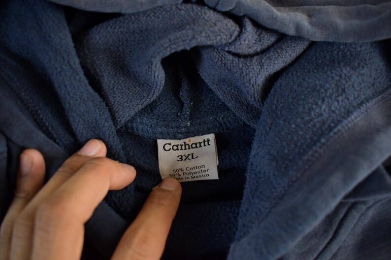 K&ouml;nnte beinhalten: Nahaufnahme eines marineblauen Carhartt-Hoodies. Das Etikett lautet "Carhartt 3XL" und gibt an, dass das Material aus 50 % Baumwolle und 50 % Polyester besteht, hergestellt in Mexiko. Der Stoff des Hoodies wirkt weich und strukturiert.